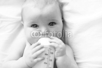 Pretty baby girl drinks water from bottle lying on bed. Child weared diaper in nursery room.