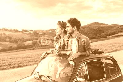 Loving couple on summer afternoon watching sunset from roof by old car around classical landscape of Tuscany, vineyards and farmland