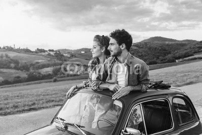 Loving couple on summer afternoon watching sunset from roof by old car around classical landscape of Tuscany, vineyards and farmland
