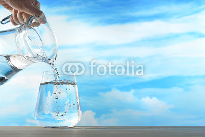 Fresh and clean drinking water being poured from jug into glass on sky background