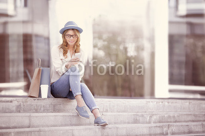 Time to communicate / Happy young woman with a disposable coffee cup and shopping bags sitting on the stairs and using her smartphone for communication via wi-fi internet in a summer park.