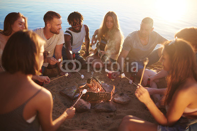 Group of restful friends roasting marshmallows while sitting by bonfire