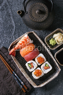 Sushi Set nigiri and sushi rolls in dark ceramic plate with soy sauce, iron teapot and chopsticks over black stone texture background. Top view with space. Japan menu