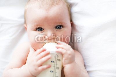 Pretty baby girl drinks water from bottle lying on bed. Child weared diaper in nursery room.