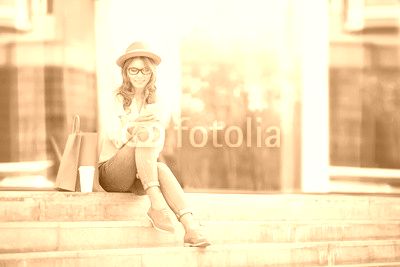 Time to communicate / Happy young woman with a disposable coffee cup and shopping bags sitting on the stairs and using her smartphone for communication via wi-fi internet in a summer park.