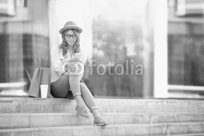 Time to communicate / Happy young woman with a disposable coffee cup and shopping bags sitting on the stairs and using her smartphone for communication via wi-fi internet in a summer park.